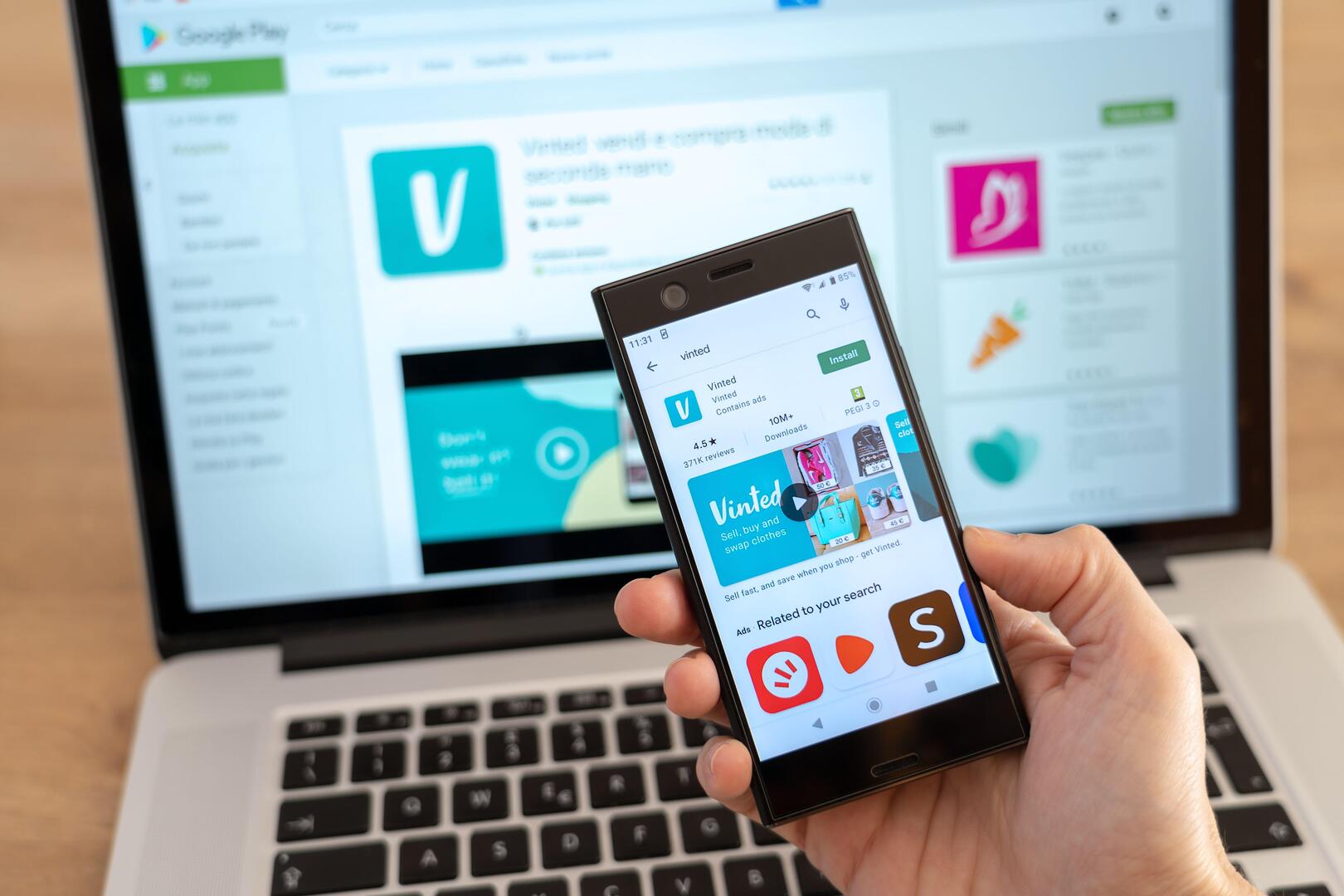 A hand holds a smartphone displaying the Vinted app on the Google Play Store, with a laptop showing the Vinted app page in the background.