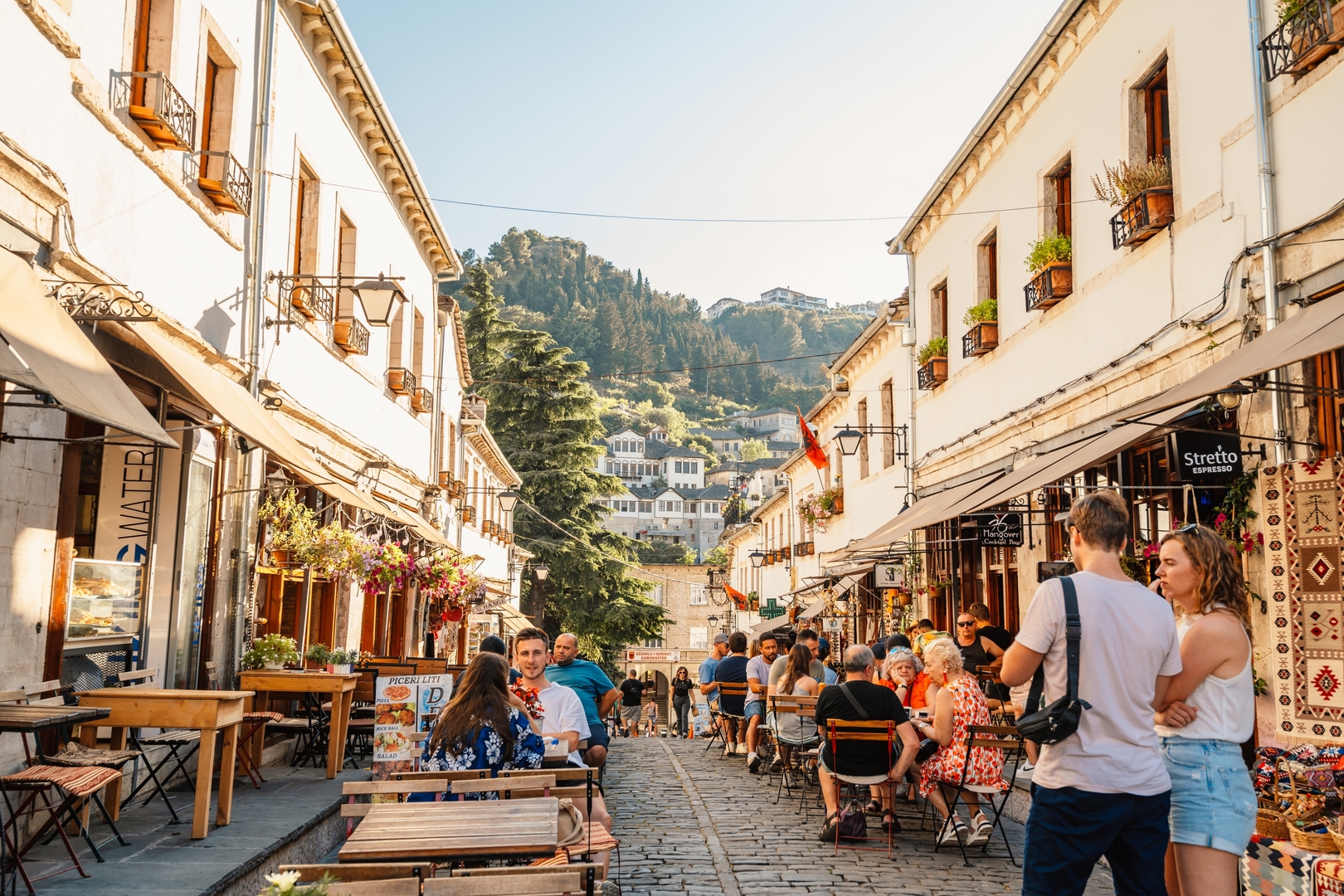 Кобblestone street in Gjirokaster, Albania, lined with outdoor cafes and souvenir shops.