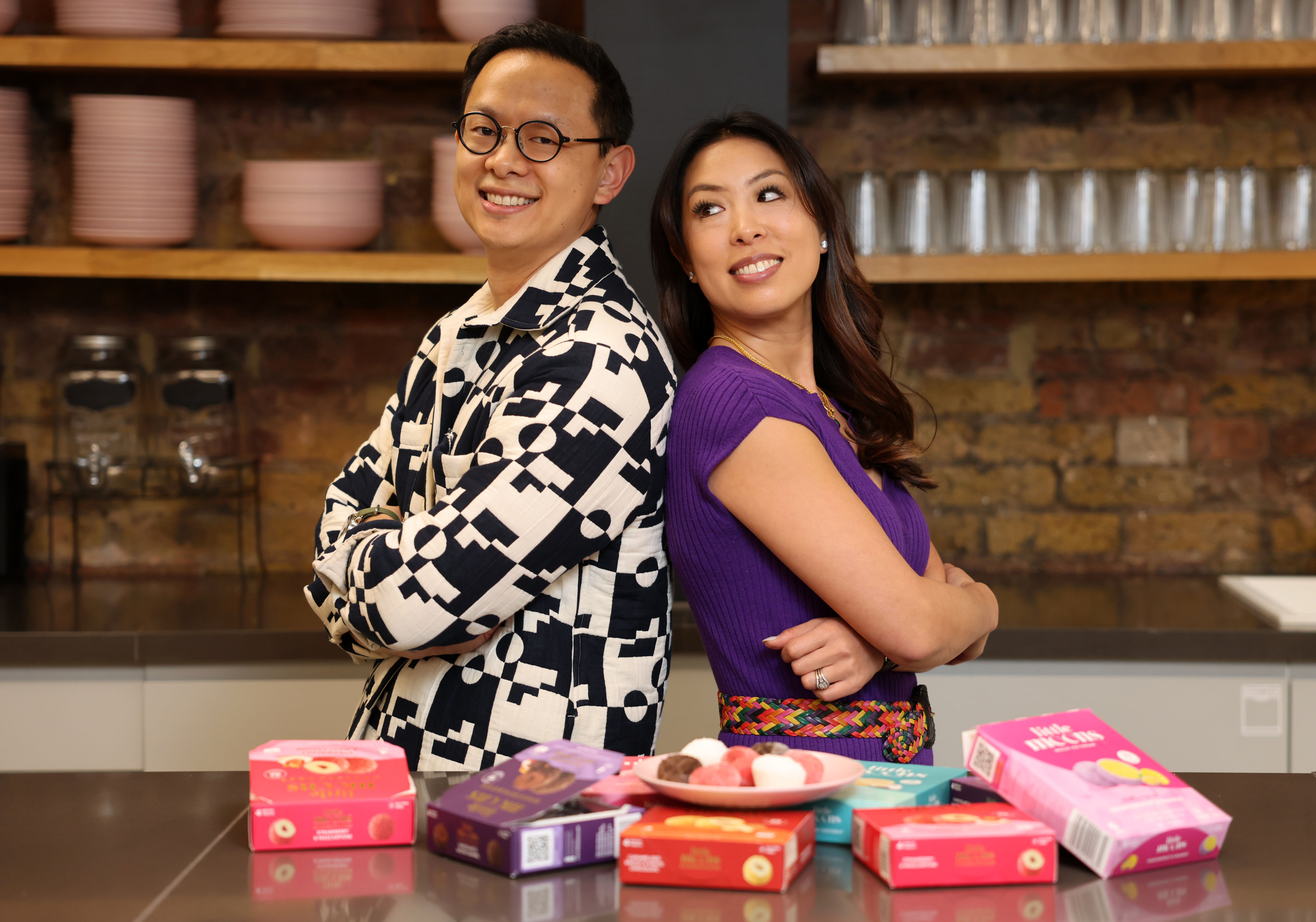 Photo of Vivien and Howard Wong, founders of Little Moons, standing back-to-back in a kitchen with boxes of their mochi ice cream.