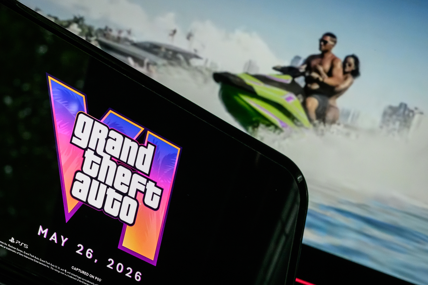 Фотоиллюстрация телефона, отображающего логотип и дату выхода Grand Theft Auto VI перед экраном с трейлером игры.